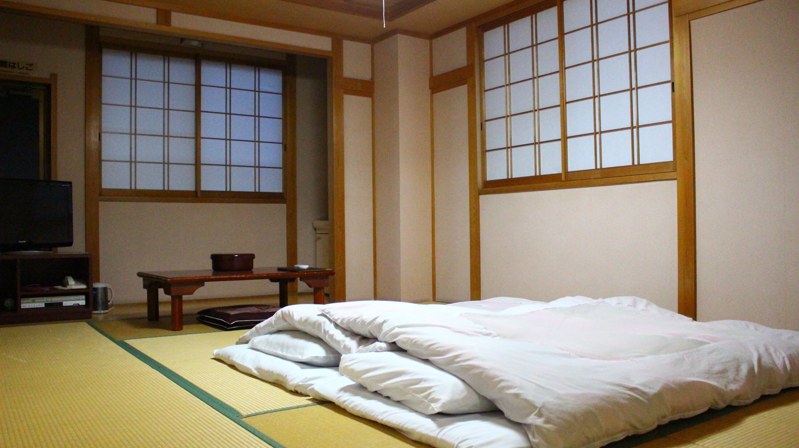 Een traditionele Japanse kamer met tatami vloerbedekking, een bed met witte beddengoed, een televisie en een klein tafeltje met een kom.