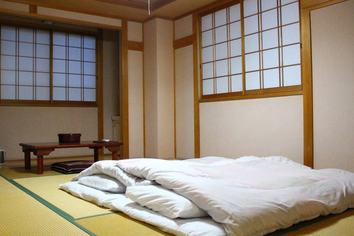 Une pièce de style japonais traditionnel, avec un sol recouvert de tatamis, un lit avec une literie blanche, une télévision et une petite table sur laquelle se trouve un bol.