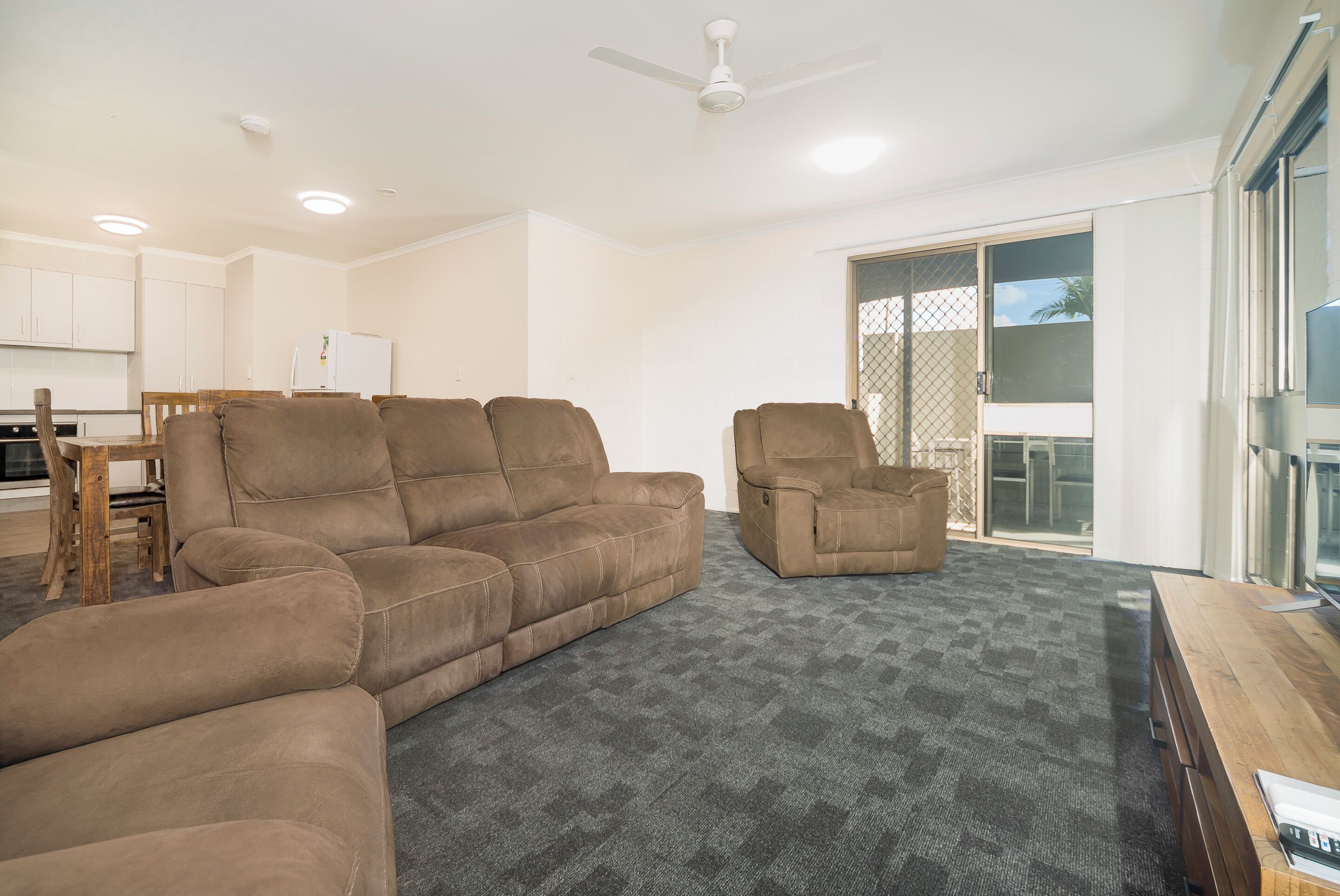 A living room with a large brown sectional sofa, a smaller armchair, and a wooden coffee table. There is a kitchen in the background with white cabinets and a refrigerator.