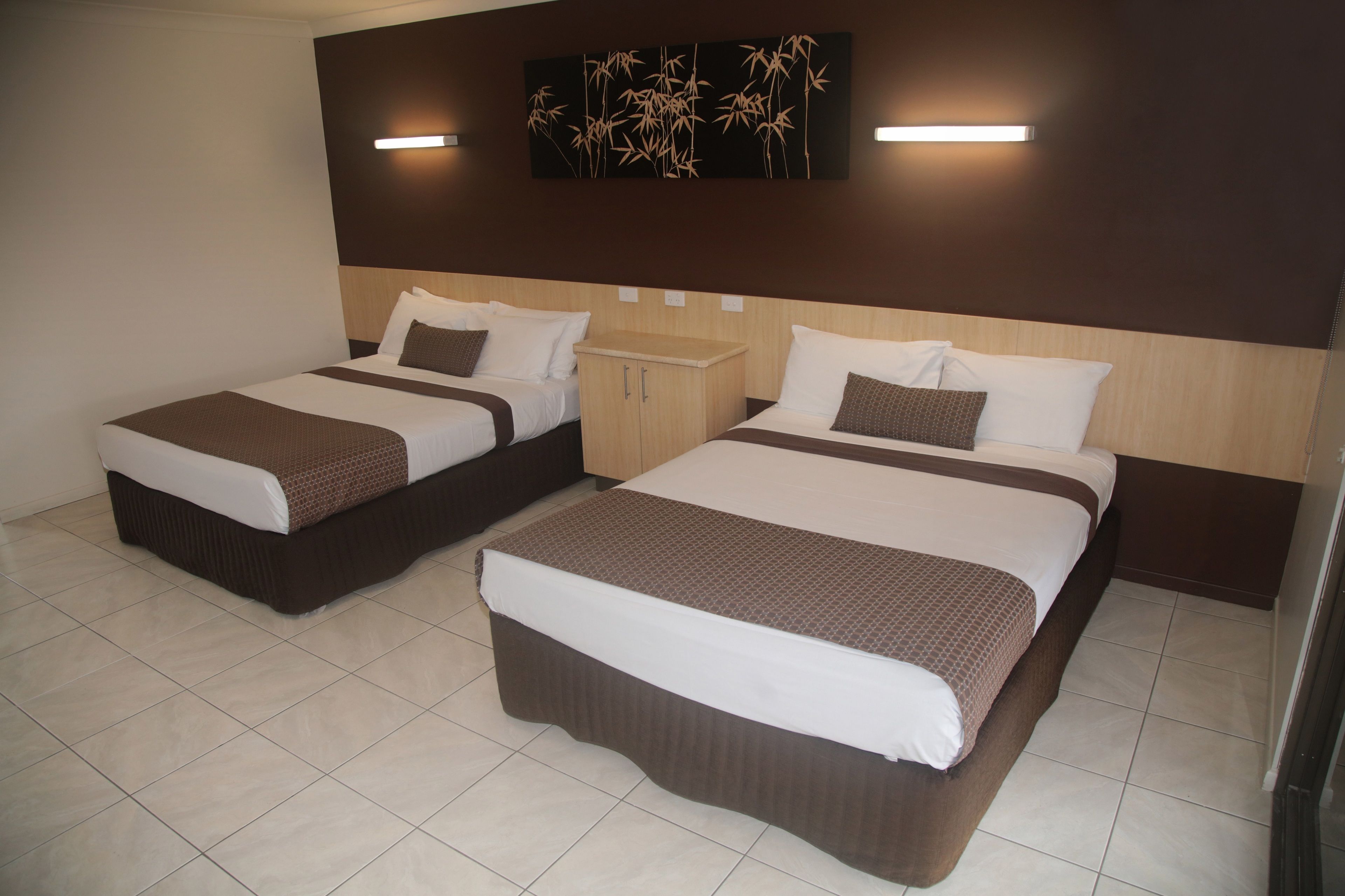 A hotel room with two beds, each with a brown bedspread and white pillows. There is a small wooden cabinet between the beds, and a wall-mounted artwork above the beds.