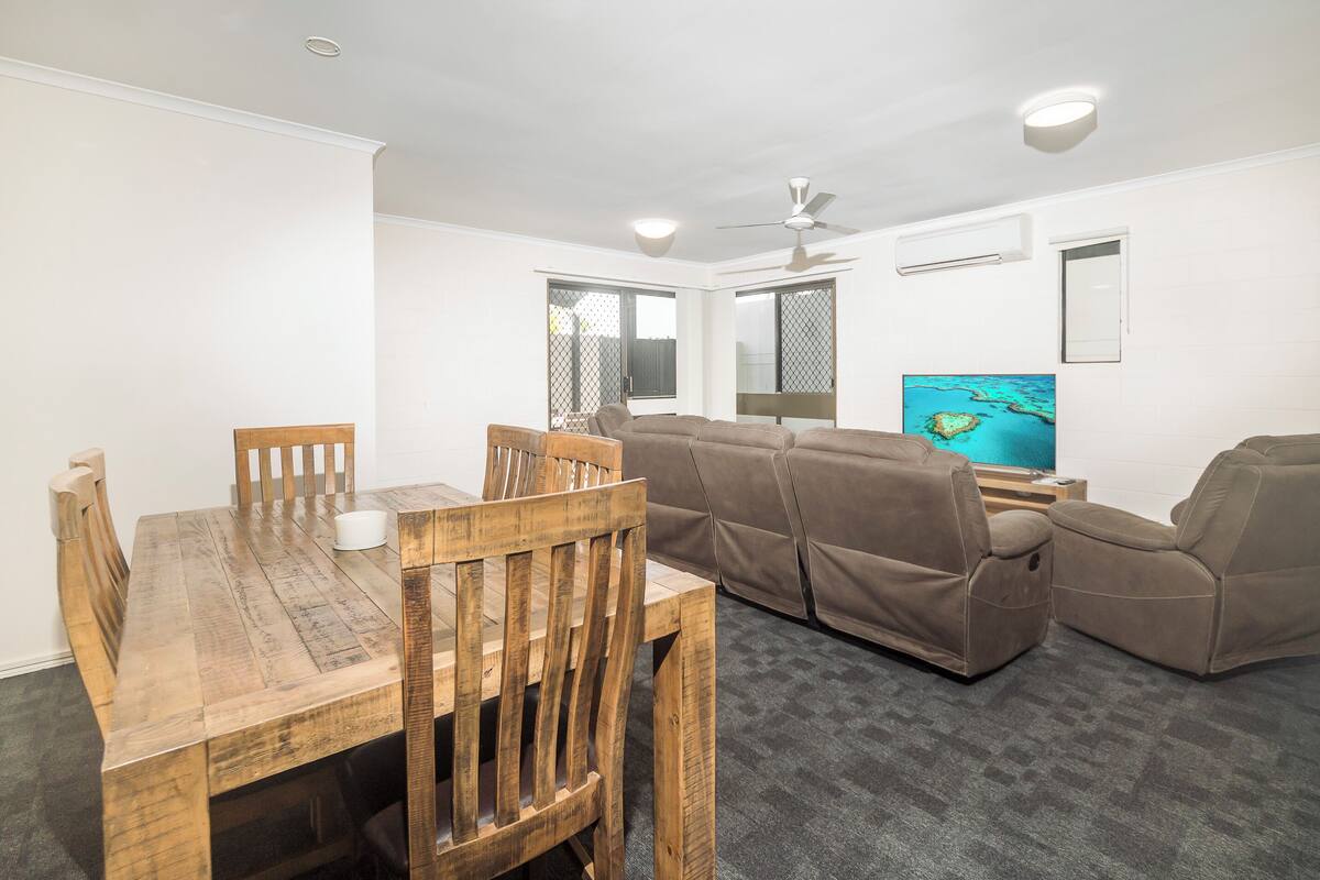 A living room with a wooden dining table, brown leather sofa, and a flat-screen TV mounted on the wall.