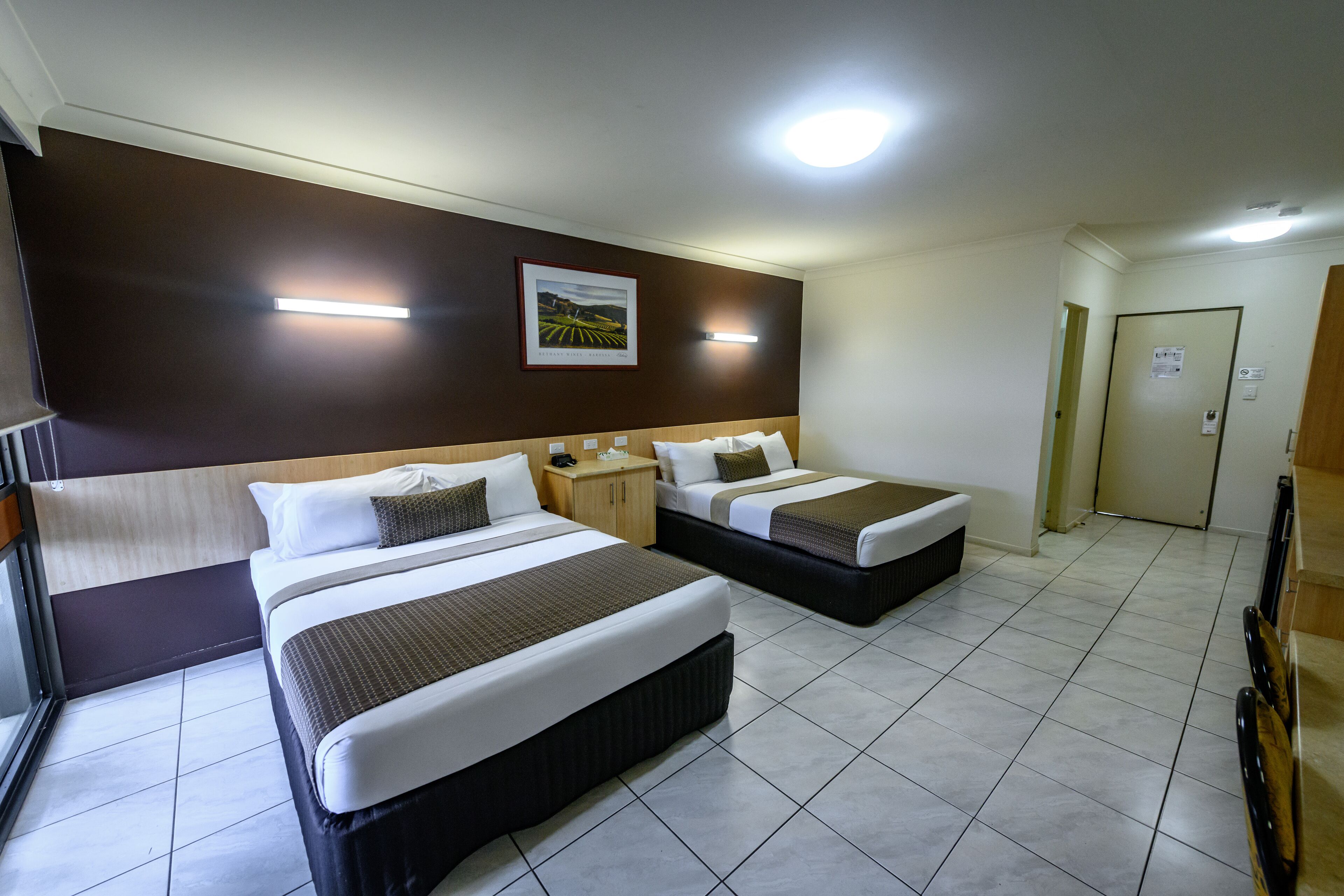 A hotel room with two beds, a nightstand, a framed picture on the wall, and a door.
