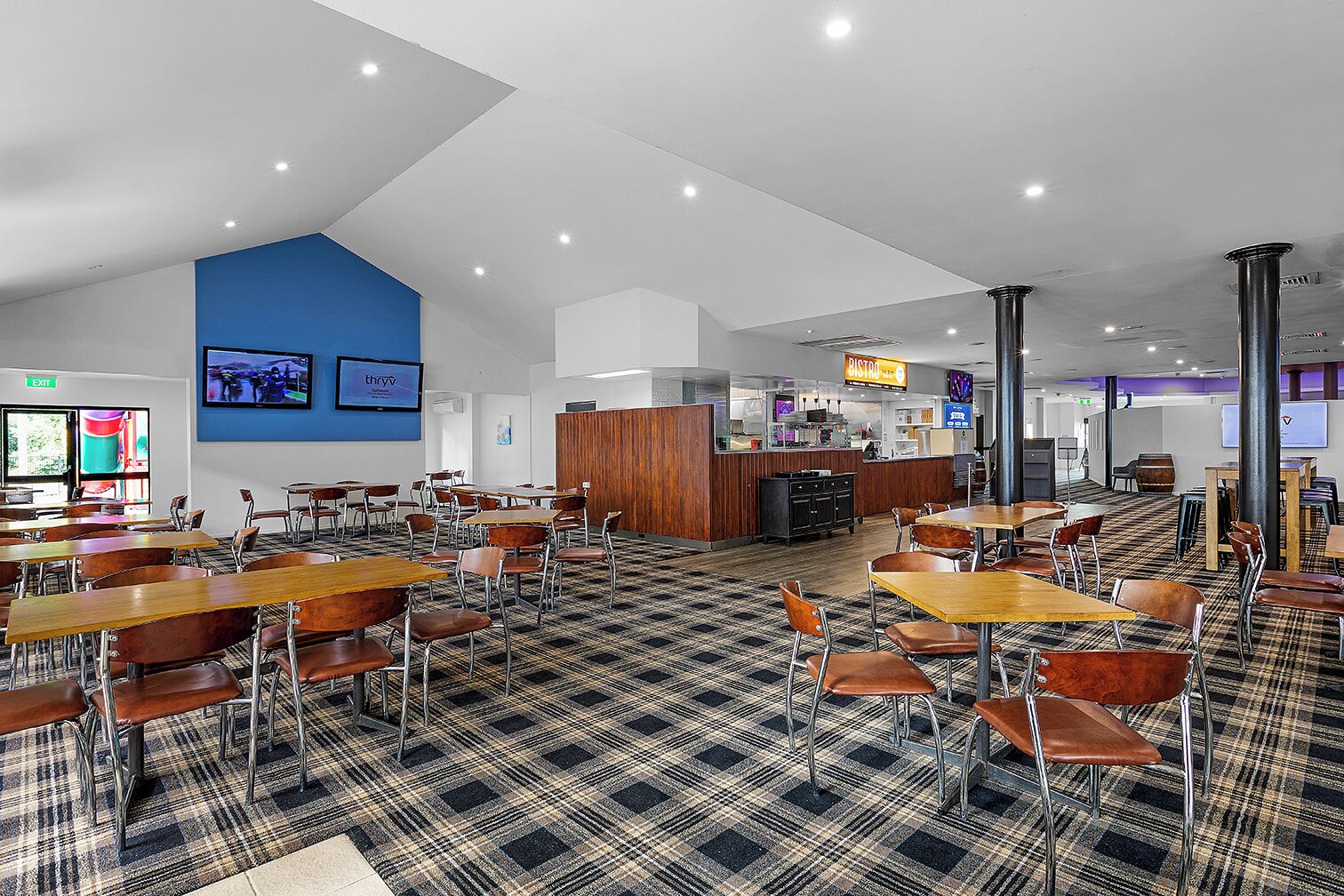 Una cafetería moderna con mesas y sillas de madera, una barra de café y una pantalla grande mostrando una presentación.