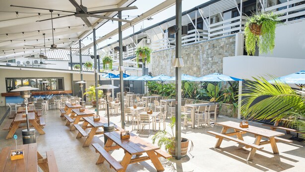 An outdoor patio area with wooden picnic tables and blue umbrellas, surrounded by potted plants and a stone wall. - Cannonvale Reef Gateway Hotel (Cannonvale)