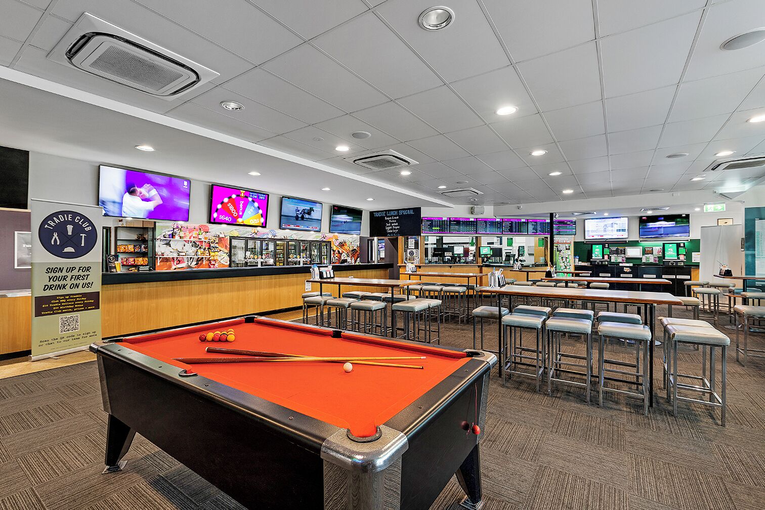 A billiards room with a pool table, bar area, and multiple screens displaying sports.