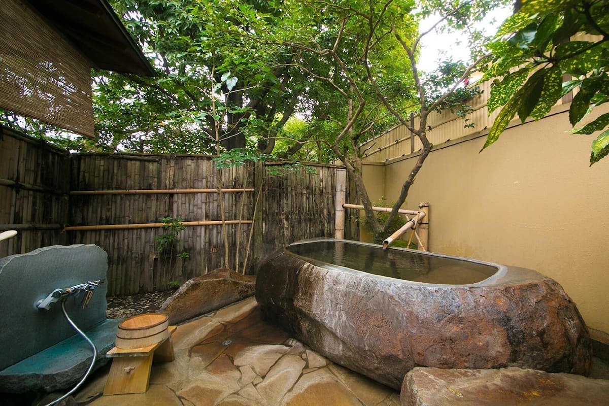 A stone hot tub in a garden with a bamboo fence and a wooden bench.