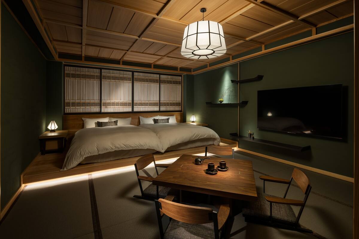 A traditional Japanese-style bedroom with a large bed, a wooden dining table with chairs, and a TV mounted on the wall.