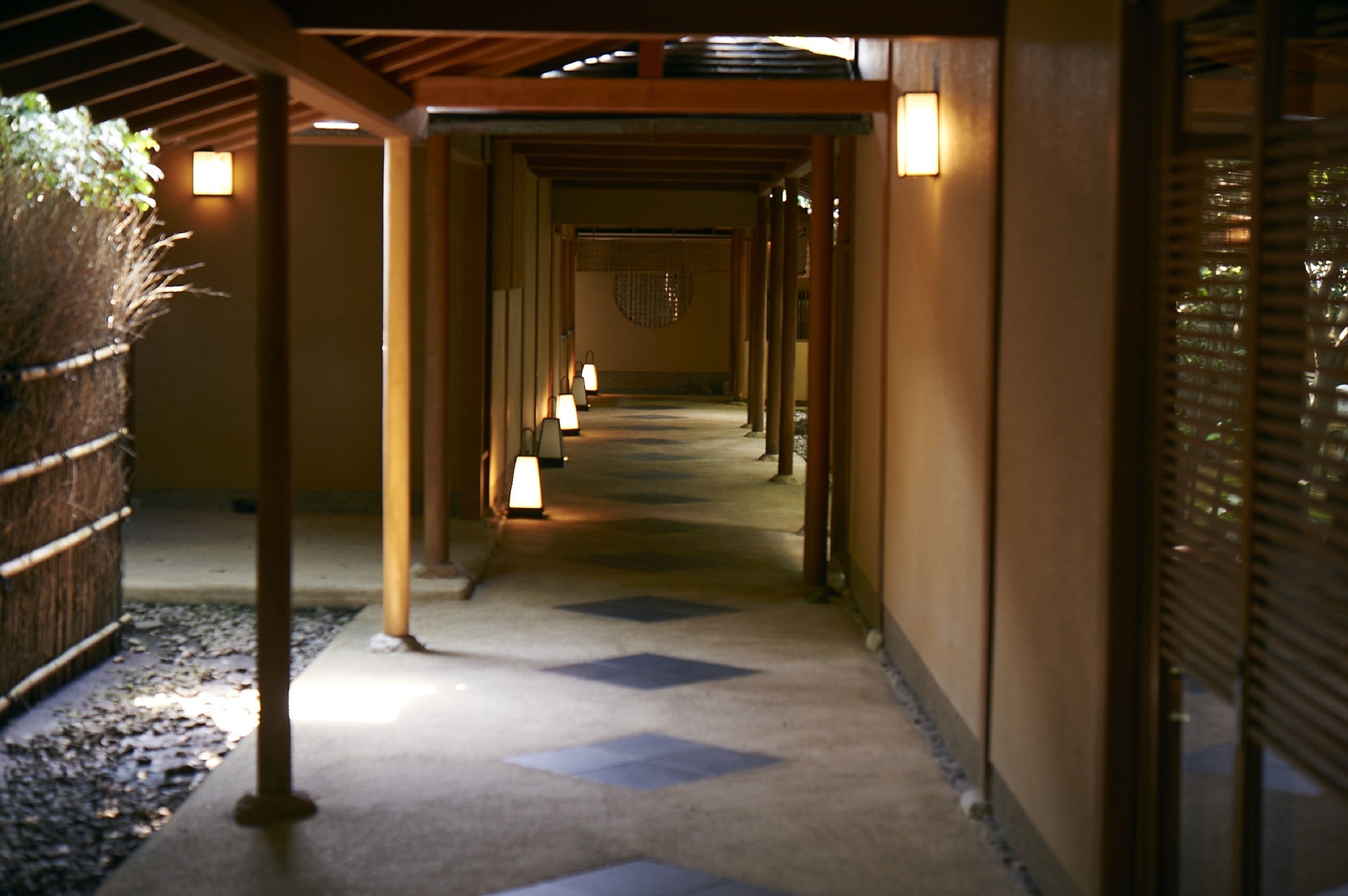 A corridor with wooden pillars, a patterned floor, and wall-mounted lights.
