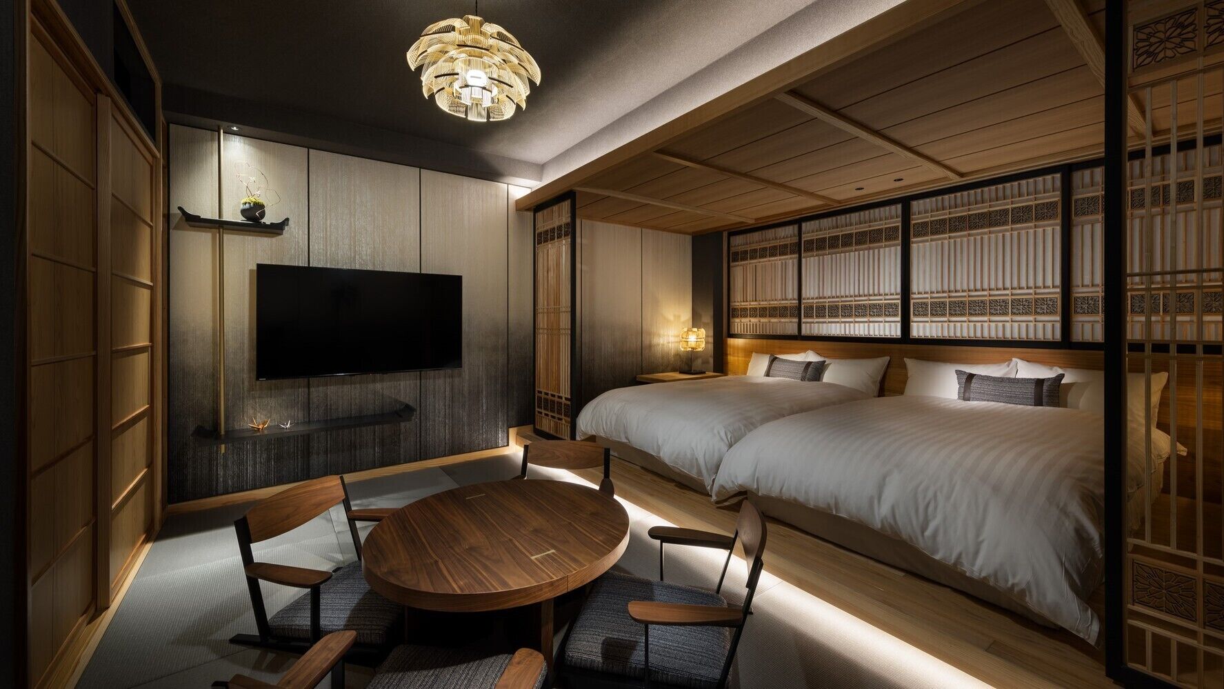 A traditional Japanese-style room with tatami flooring, a round wooden table, two beds with white bedding, and a television mounted on the wall.