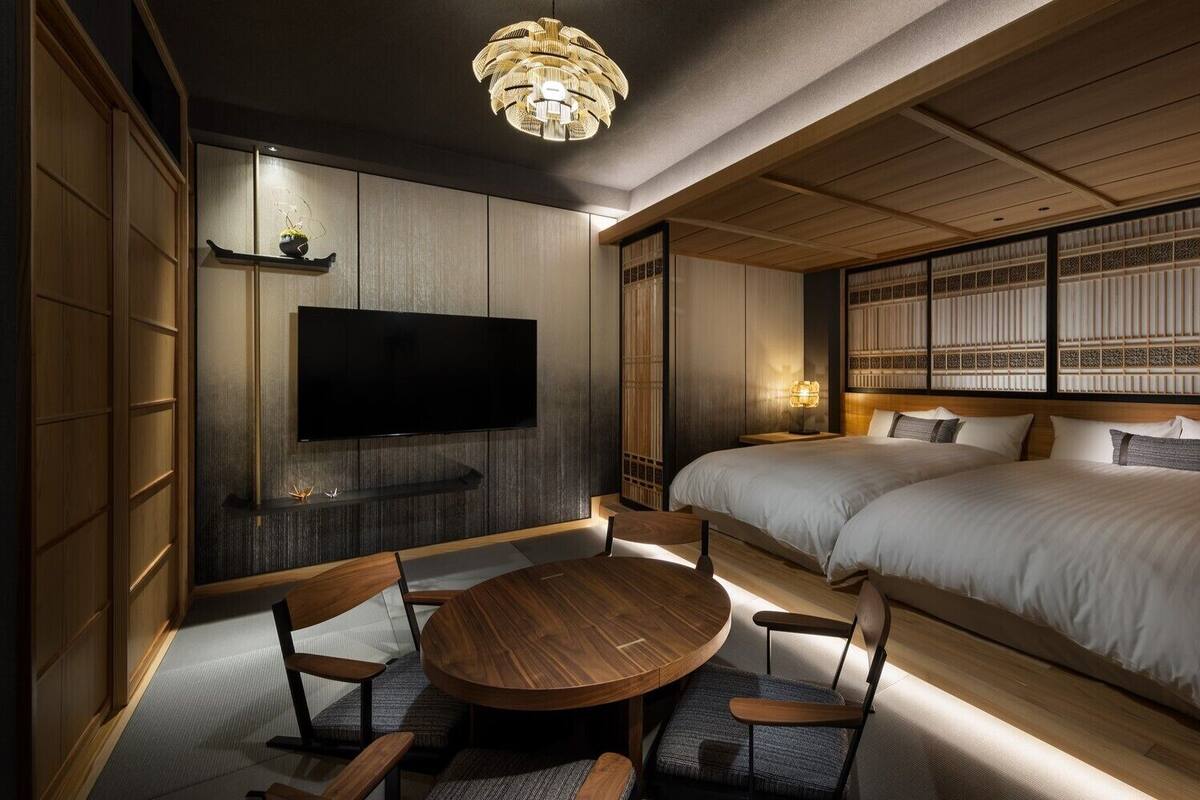 A traditional Japanese-style room with tatami flooring, a round wooden table, two beds with white bedding, and a television mounted on the wall.