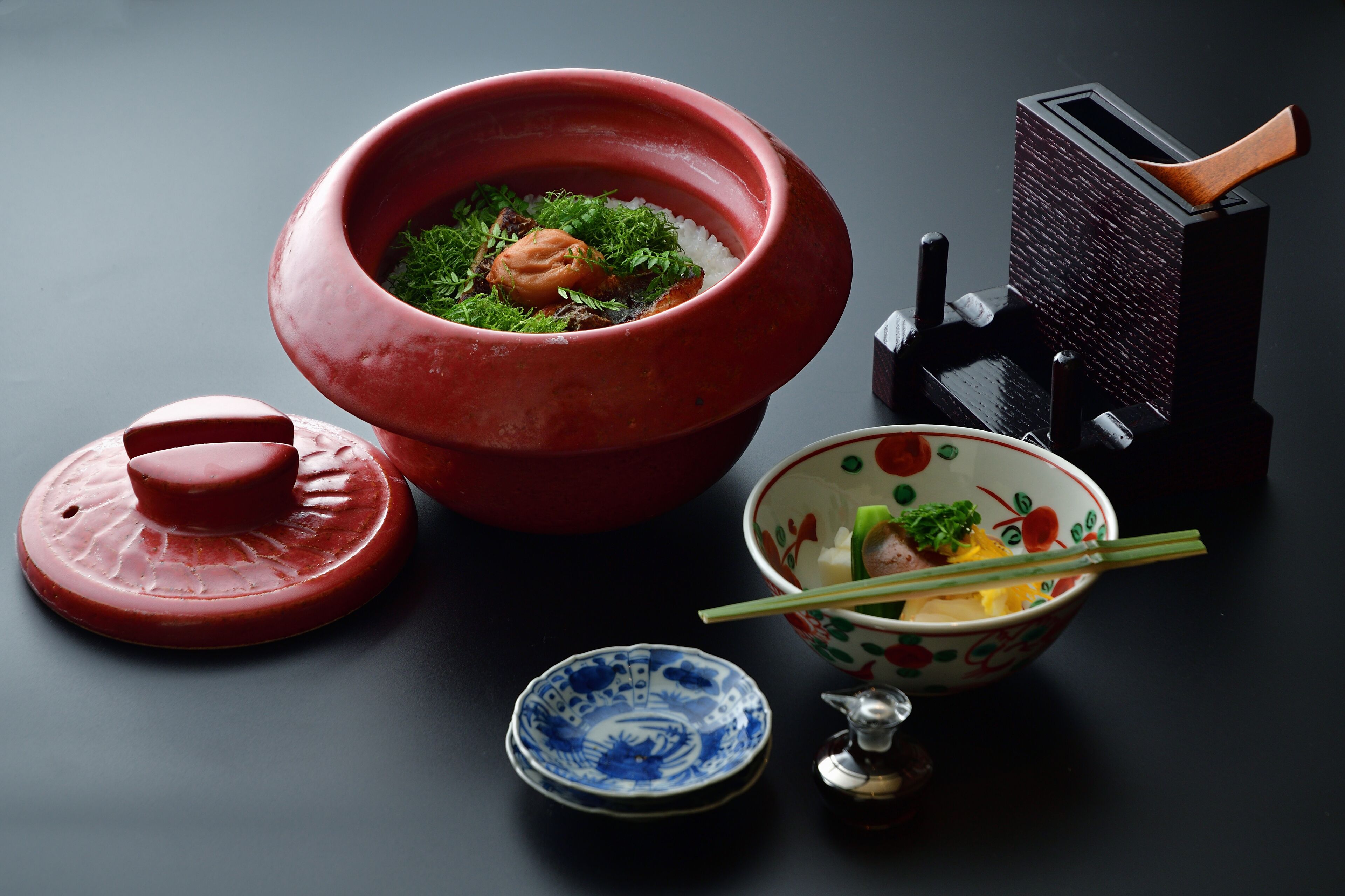 A traditional Japanese tea set with a red tea bowl, a small blue and white tea bowl, a tea scoop, and a wooden brush.