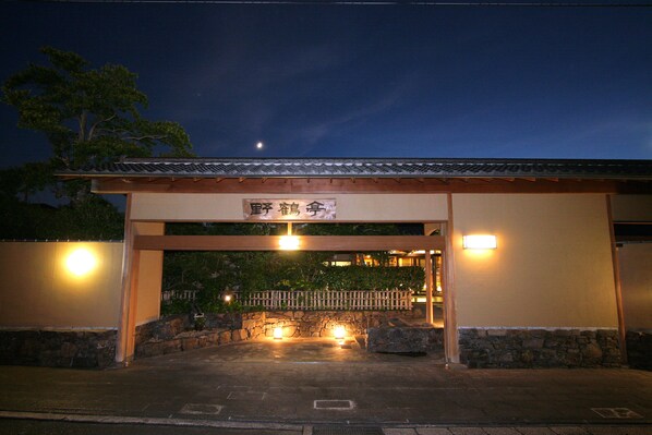 Entrada de un edificio tradicional japonés de noche, con un letrero, pared de piedra y valla de madera.