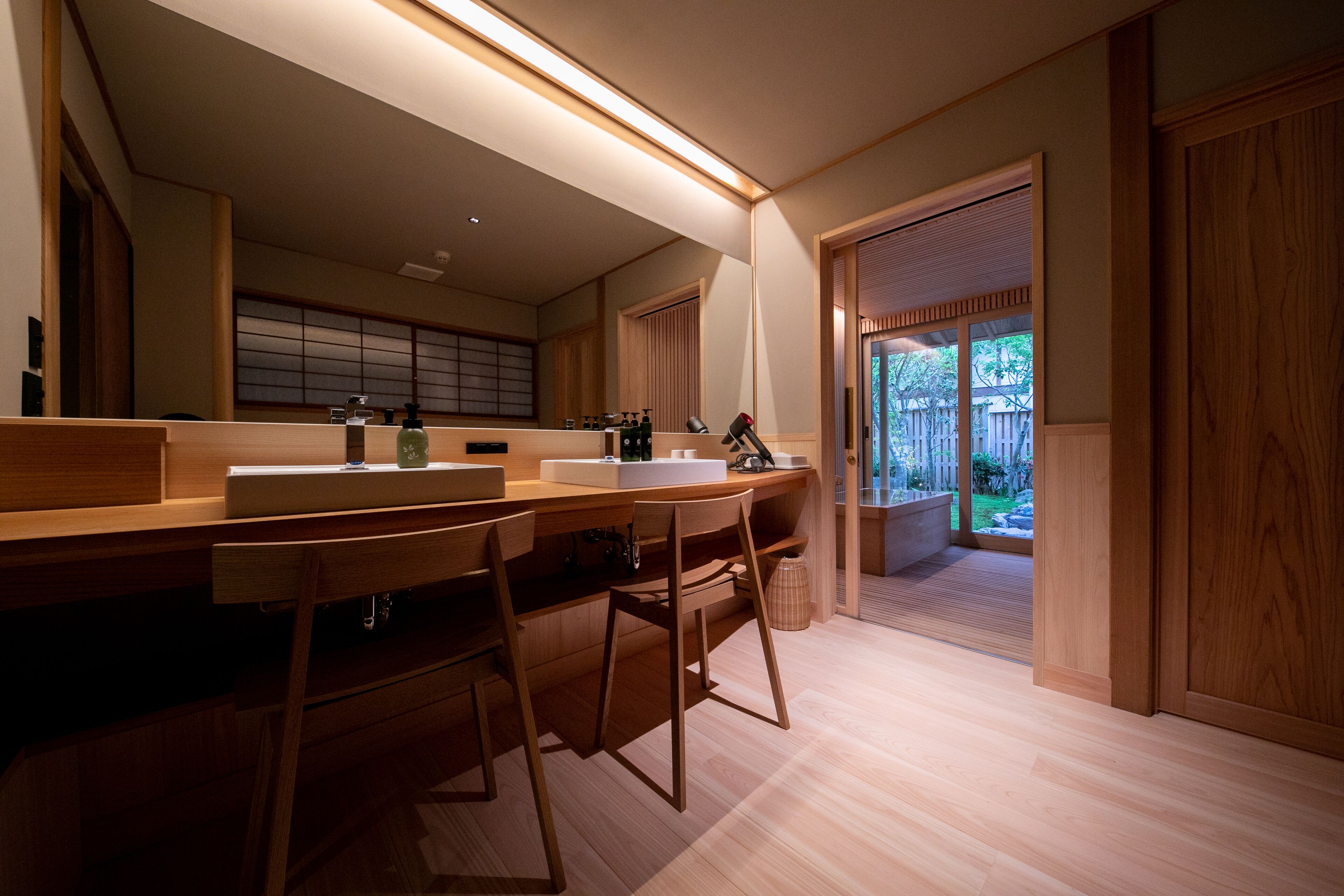 Une salle de bain moderne avec des meubles en bois, un grand miroir et une vue sur un jardin, visible à travers une porte ouverte.