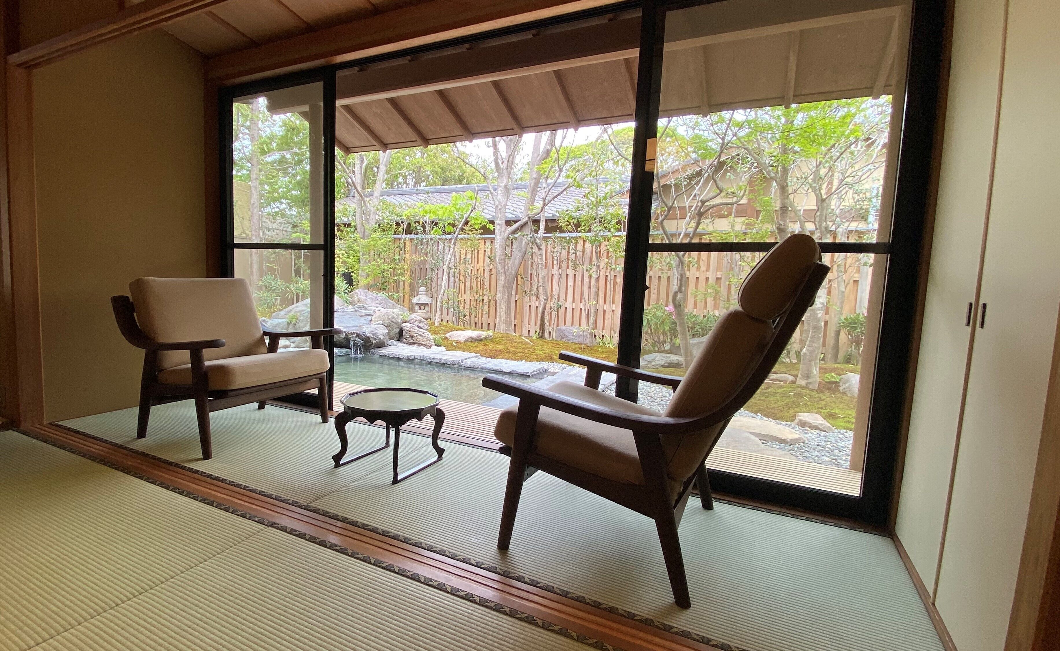 A room with tatami mat flooring, a sliding glass door leading to a garden with a koi pond, two wooden chairs, and a small table.