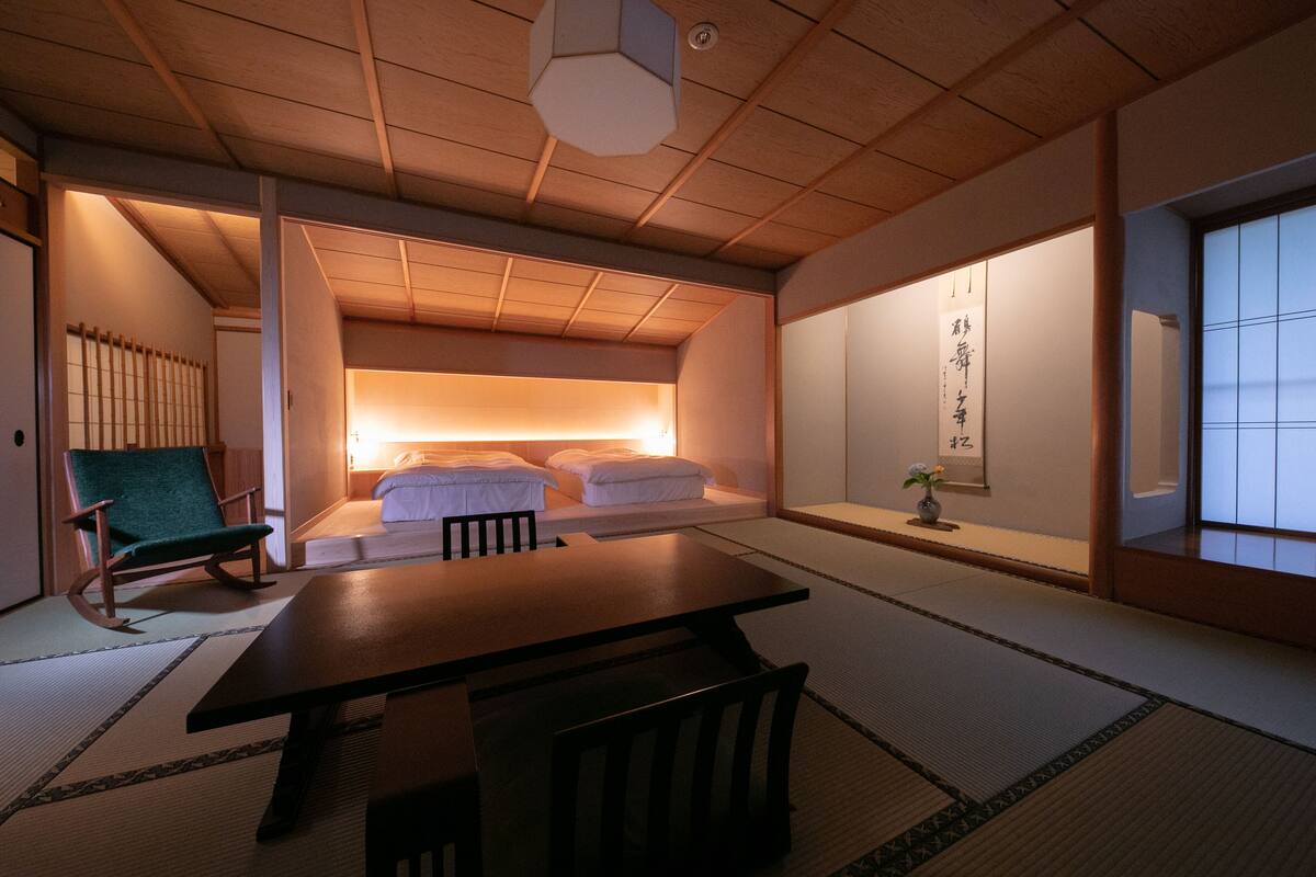 A traditional Japanese room with tatami flooring, a low table, a green chair, and a futon bed with white bedding.