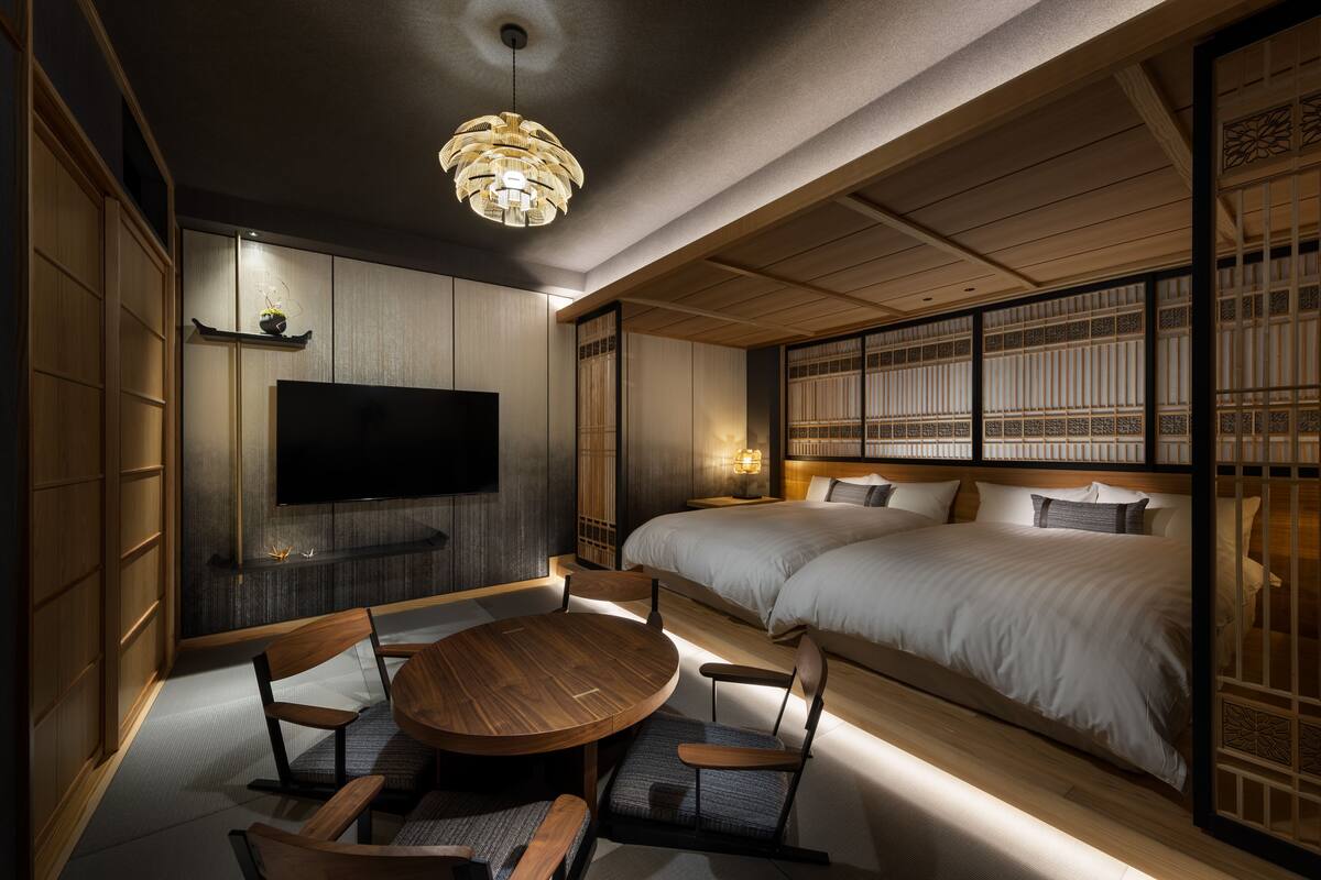 A traditional Japanese-style room with tatami flooring, a low wooden bed, a television, and a round wooden table with chairs.