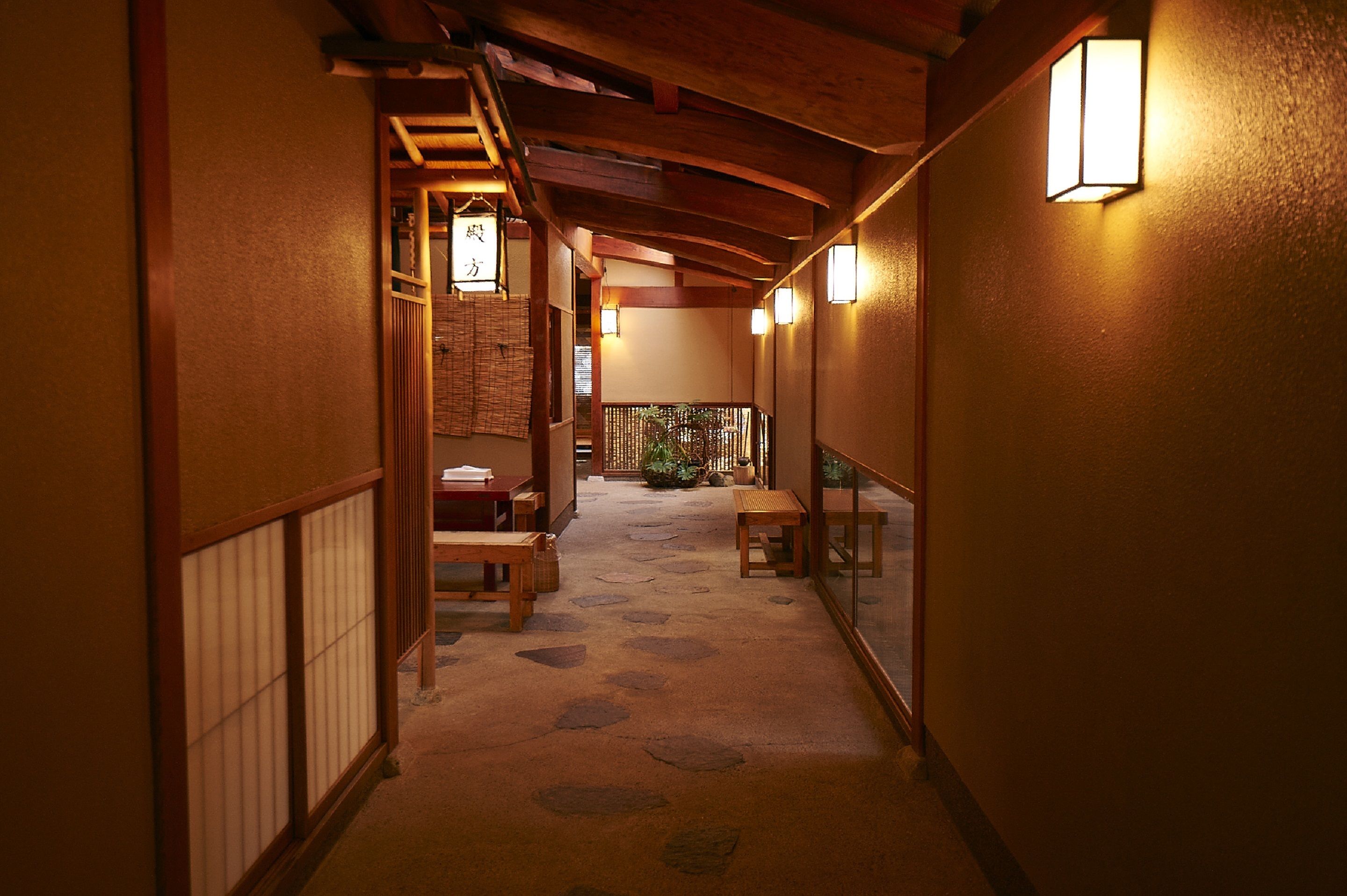 A narrow hallway with wooden beams and tatami flooring, leading to a room with a sliding door and a small table.