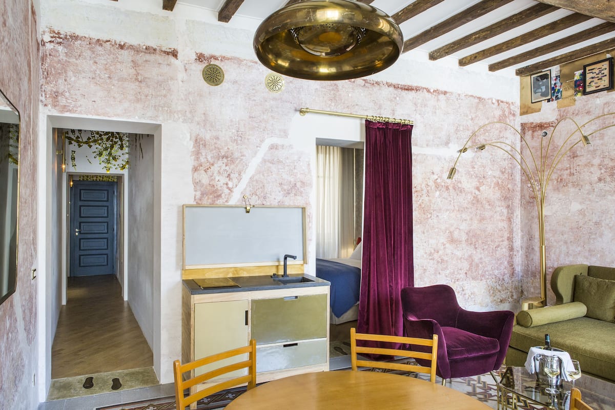 A room with a wooden dining table, a purple armchair, a green sofa, a small kitchenette, and a ceiling light fixture.