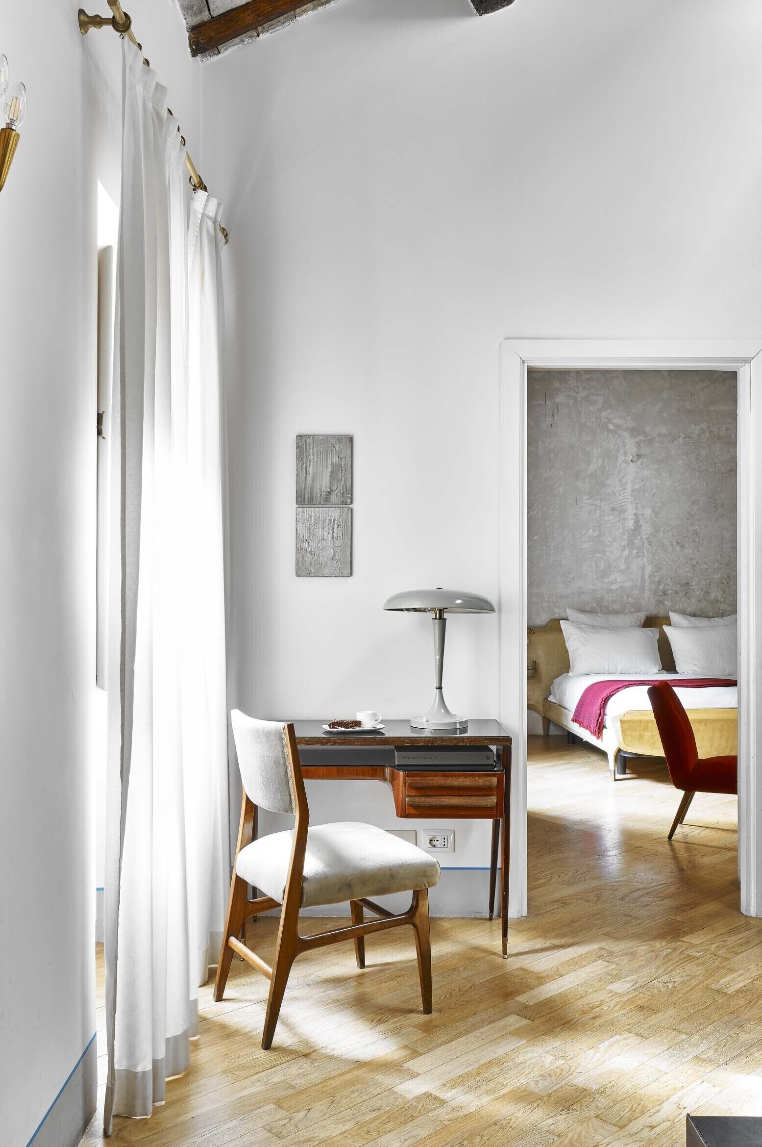 Una stanza con una scrivania in legno, una sedia, una lampada, un letto con una poltrona rossa e un muro in cemento.