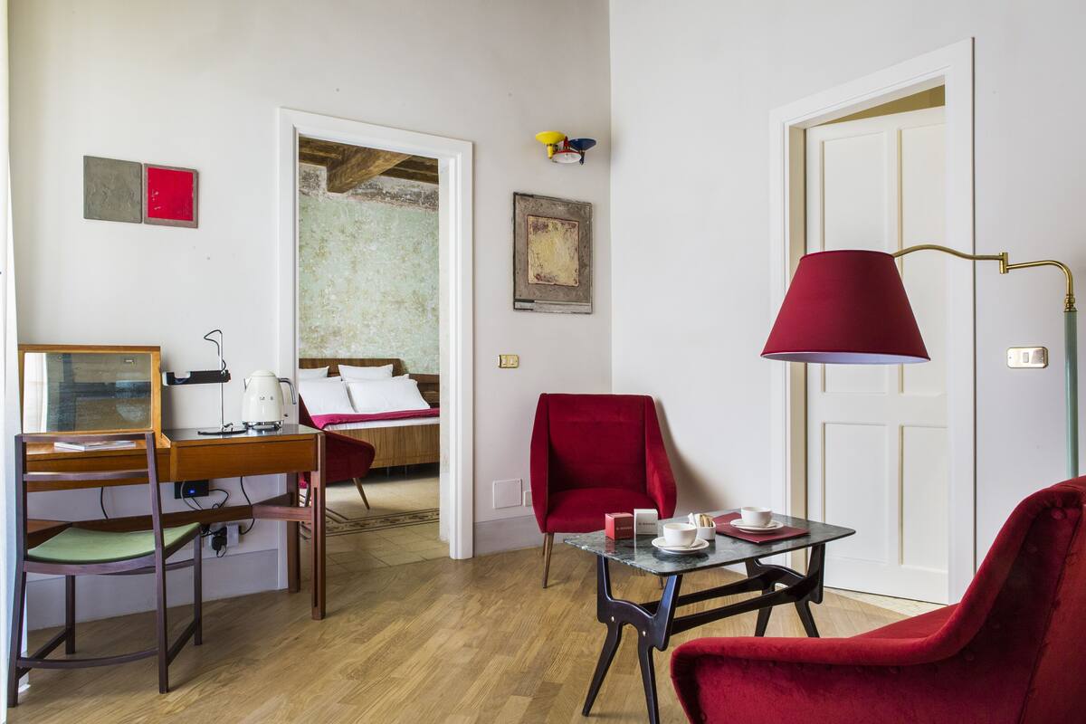 A compact hotel room with a wooden desk, a red armchair, a small table with a lamp, and a bed with white bedding.