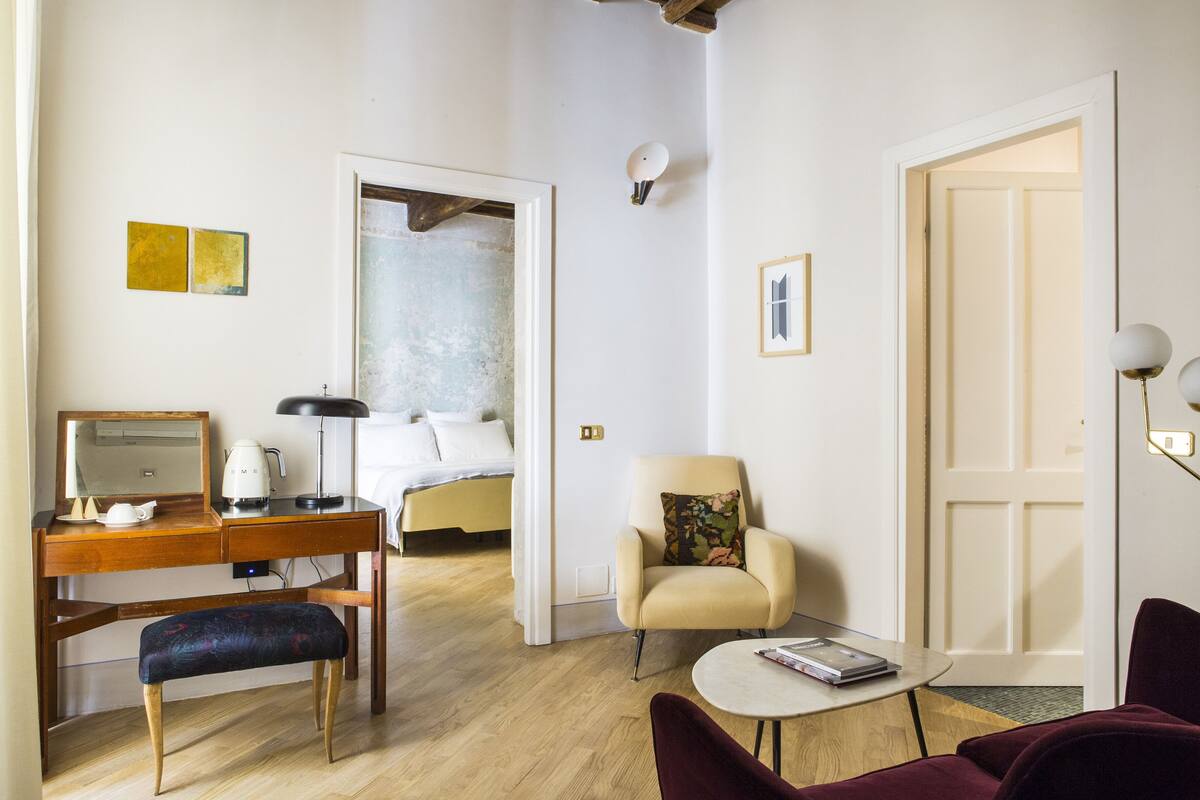 A modern hotel room with a wooden desk, a comfortable armchair, a round coffee table, and a bed with a view of the city.