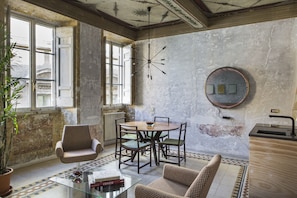 A room with a large window, a round table with chairs, a potted plant, and a glass coffee table with books.