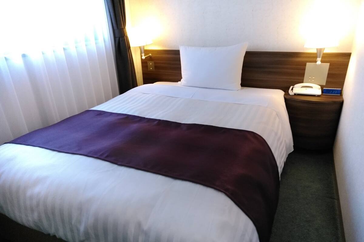 Een hotelkamer met een net opgemaakt bed, een bedlamp, een klok en een telefoon op het nachtkastje.