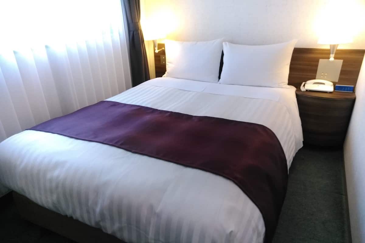 A hotel room with a bed, a nightstand with a phone, and a window with blinds.