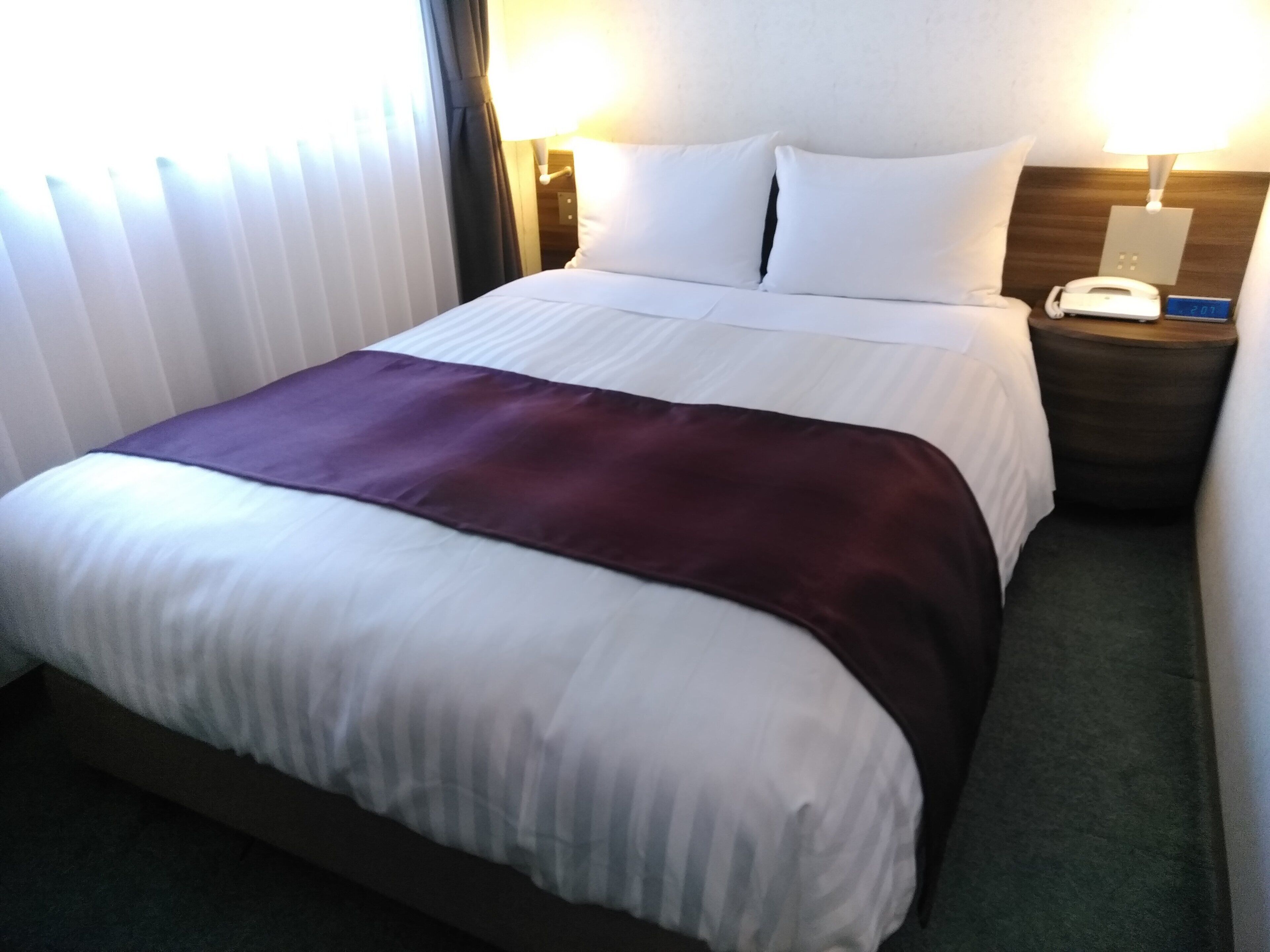 Una habitación de hotel con una cama, una mesita de noche con teléfono y una lámpara de pared.