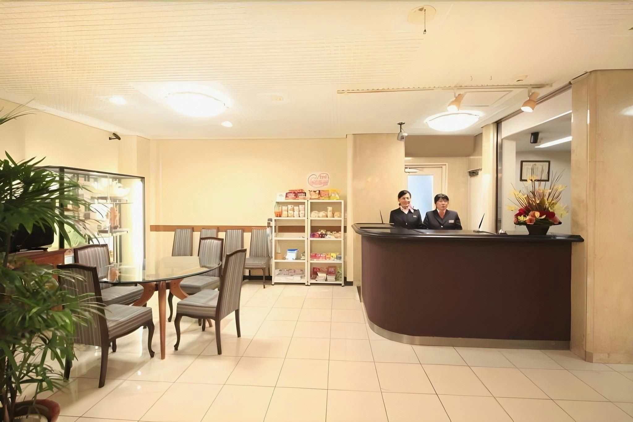 A hotel reception area with a reception desk, chairs, a small dining table, and a shelf with various items.