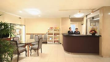 A hotel reception area with a reception desk, chairs, a small dining table, and a shelf with various items.