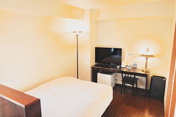 A hotel room with a bed, a desk with a television, a lamp, a chair, and a small refrigerator.
