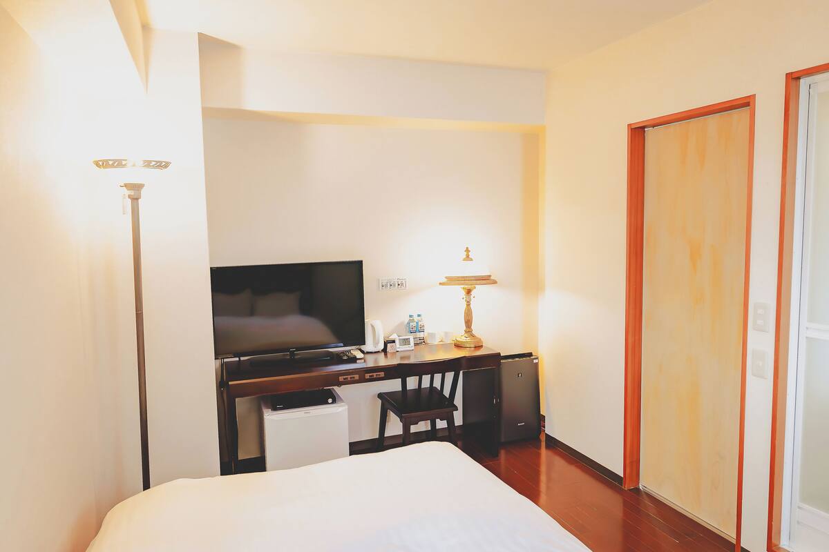 A hotel room with a bed, a desk with a television, a chair, a lamp, and a wardrobe.