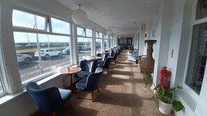 Un pasillo de hotel con filas de sillas y mesas azules, ventanas amplias que dan vistas a coches aparcados y un jarrón de mimbre a la derecha.