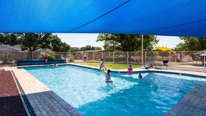 Une piscine rectangulaire surmontée d’une voile d’ombrage bleue, entourée d’une clôture et d’arbres.