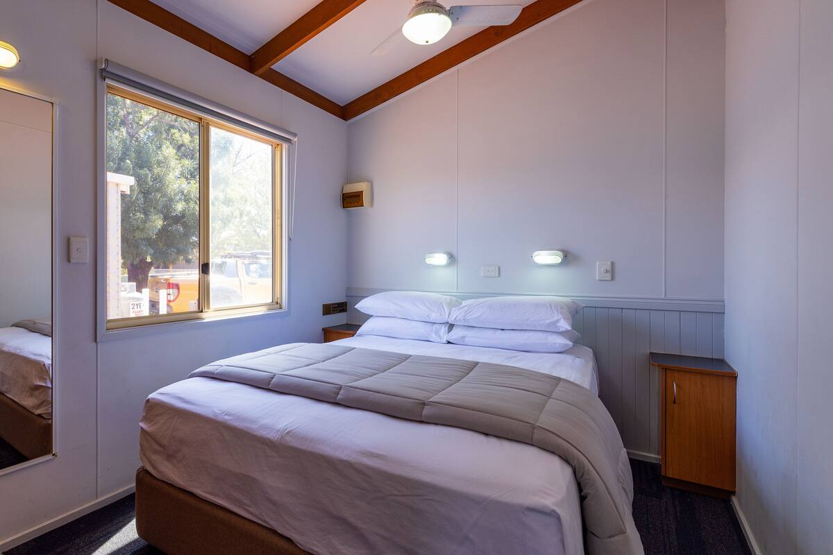Quarto com uma cama grande, mesa de cabeceira, armário de madeira e janela com vista para árvores.