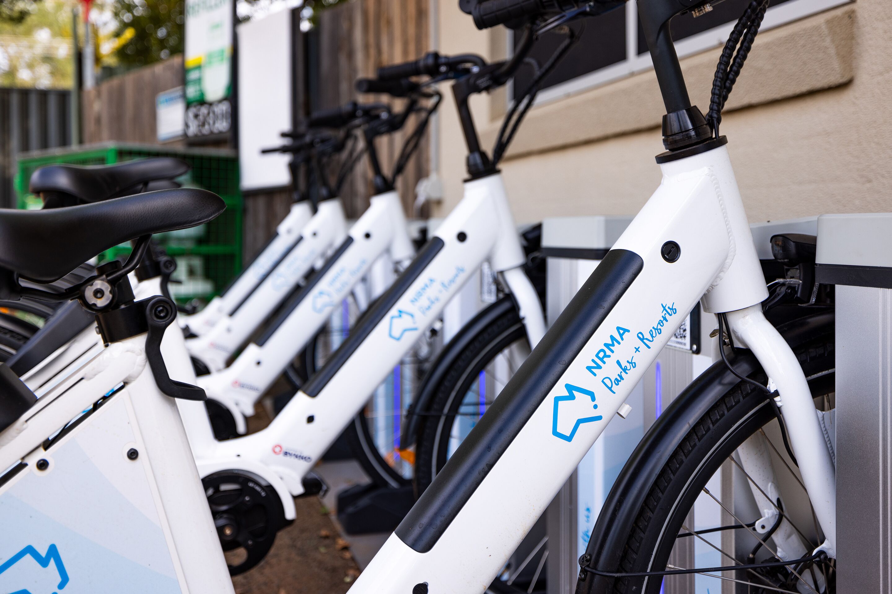 Une rangée de vélos électriques blancs, ornés de logos bleus, est stationnée devant un immeuble.