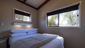 Une chambre à coucher avec un lit, deux fenêtres équipées de stores, une table de chevet et une commode.