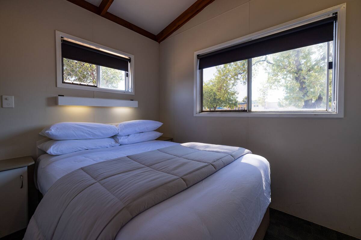 Quarto com cama, duas janelas com persianas, mesa de cabeceira e criado-mudo.