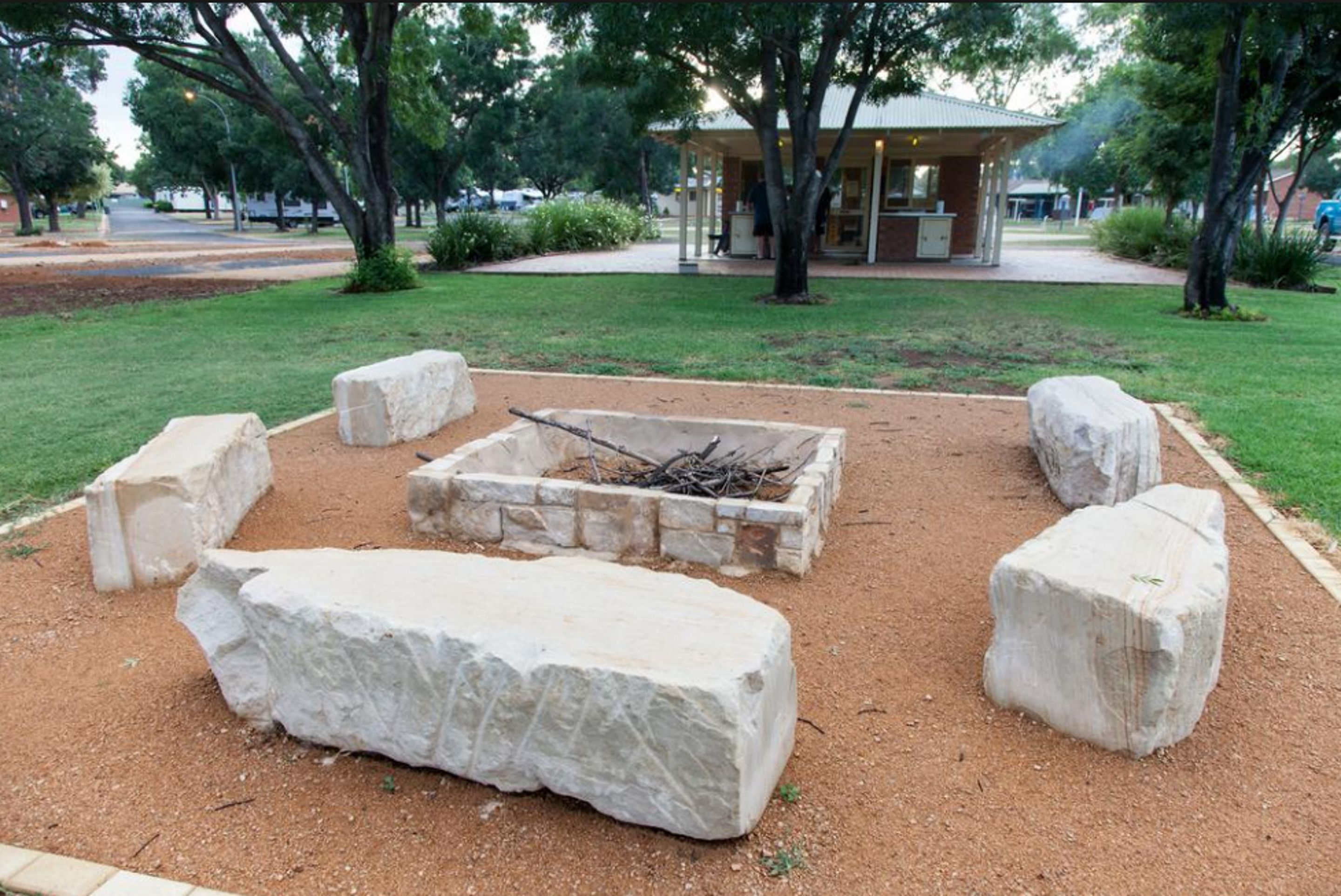 A fire pit surrounded by large white stones in a landscaped area with trees and a building in the background.