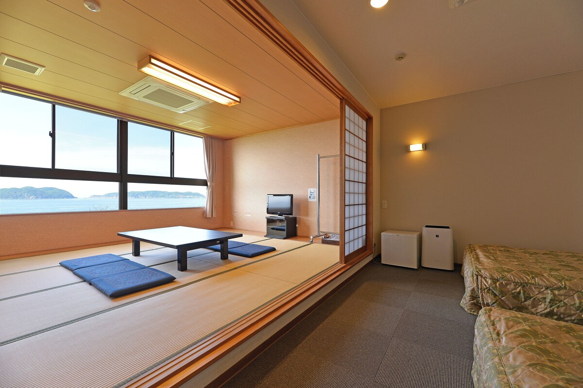 Una habitación con suelo de tatami, una cama futón, una mesa con cojines, un televisor y vistas al exterior.