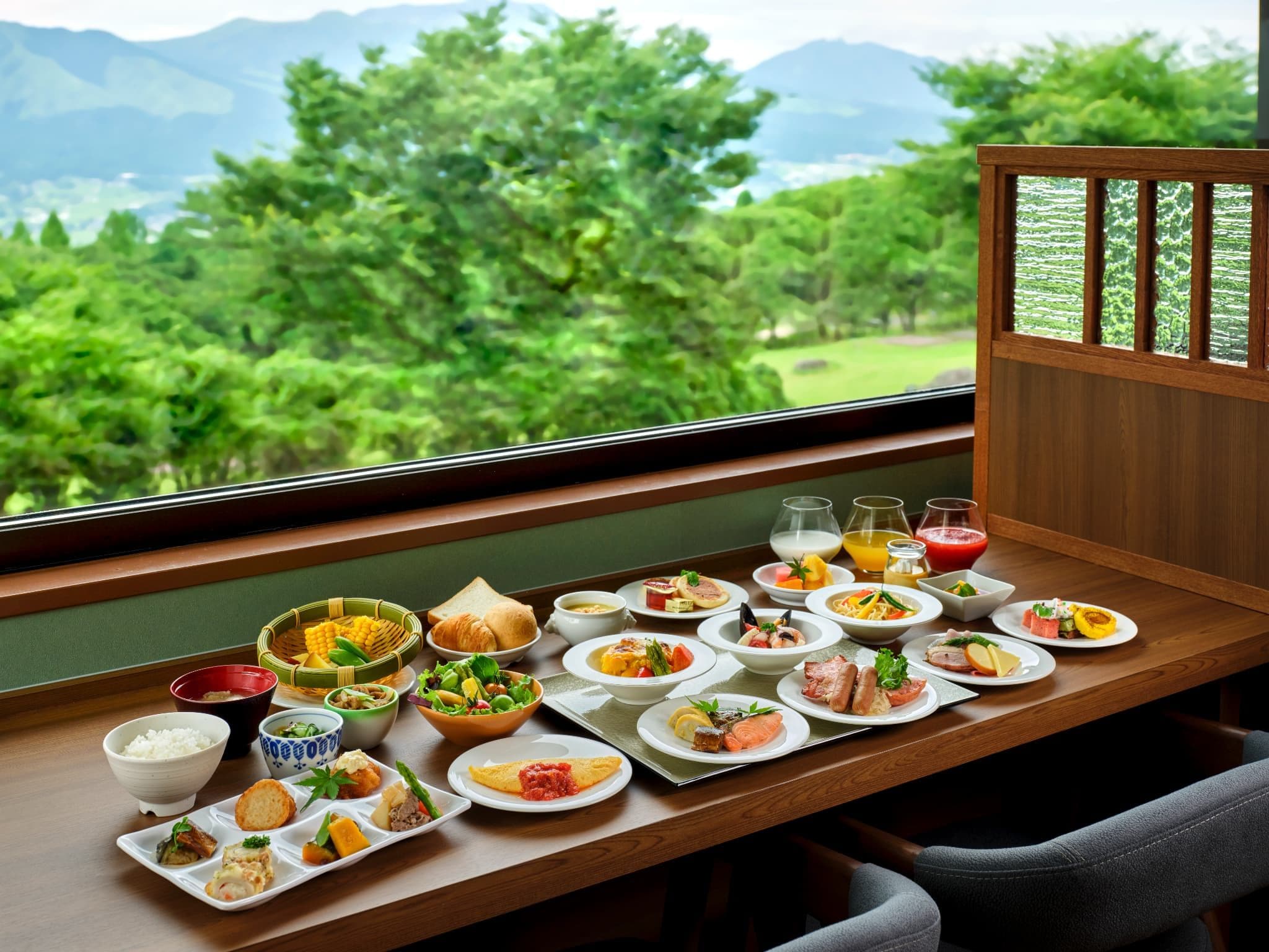 Una mesa de comedor con diversos platos y bebidas, frente a una ventana con vistas a vegetación y montañas.