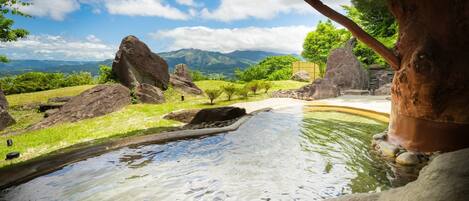 A stone water feature with a wooden structure overhead, surrounded by rocks and greenery, with mountains in the background.