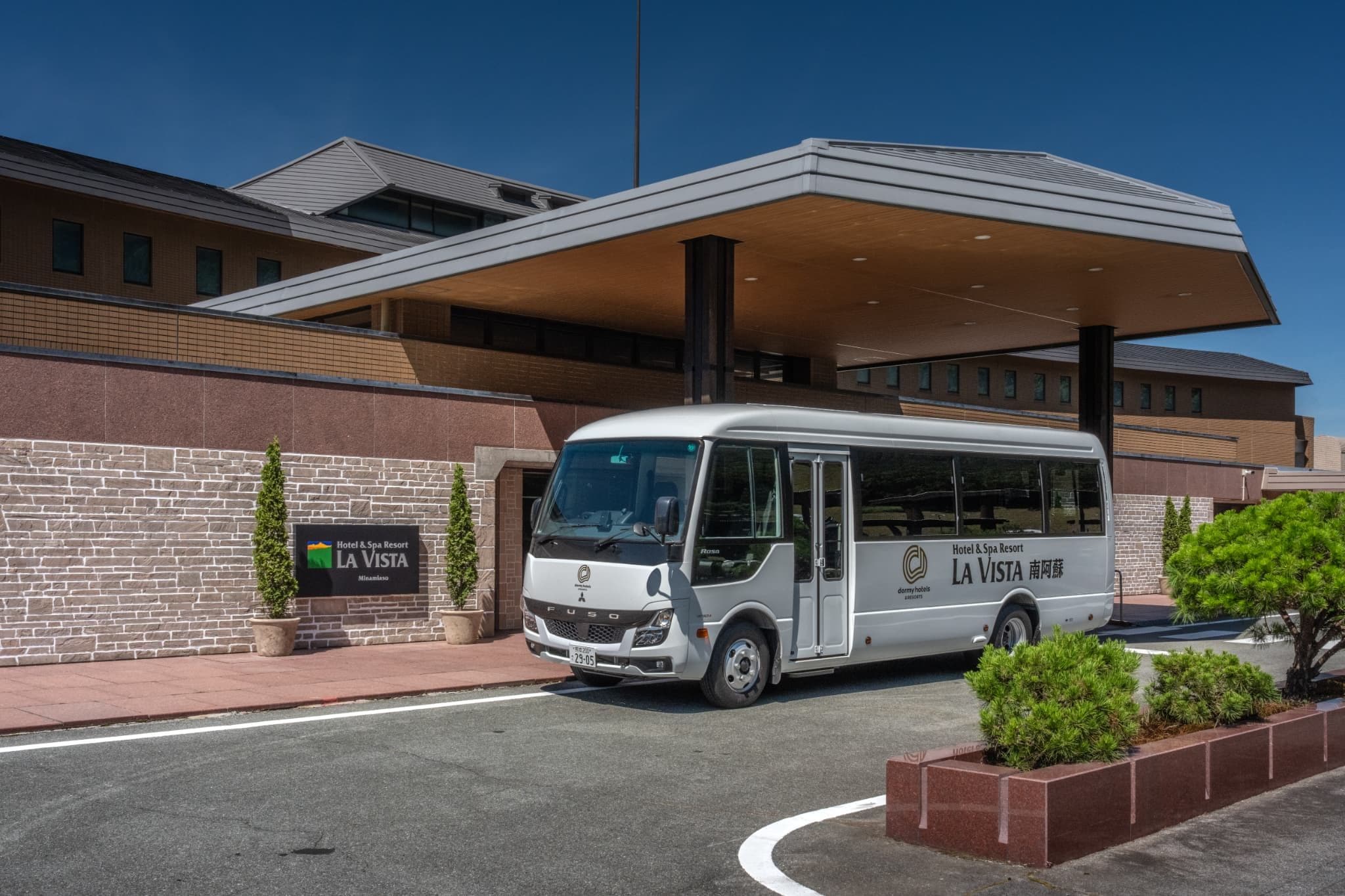 Un autobús blanco estacionado frente a un hotel de estilo arquitectónico moderno, con fachada de piedra y un cielo azul despejado.