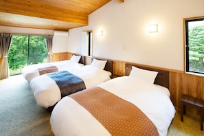 Une chambre d’hôtel avec trois lits, des poutres en bois au plafond, une fenêtre donnant sur un paysage verdoyant et un climatiseur fixé au mur.