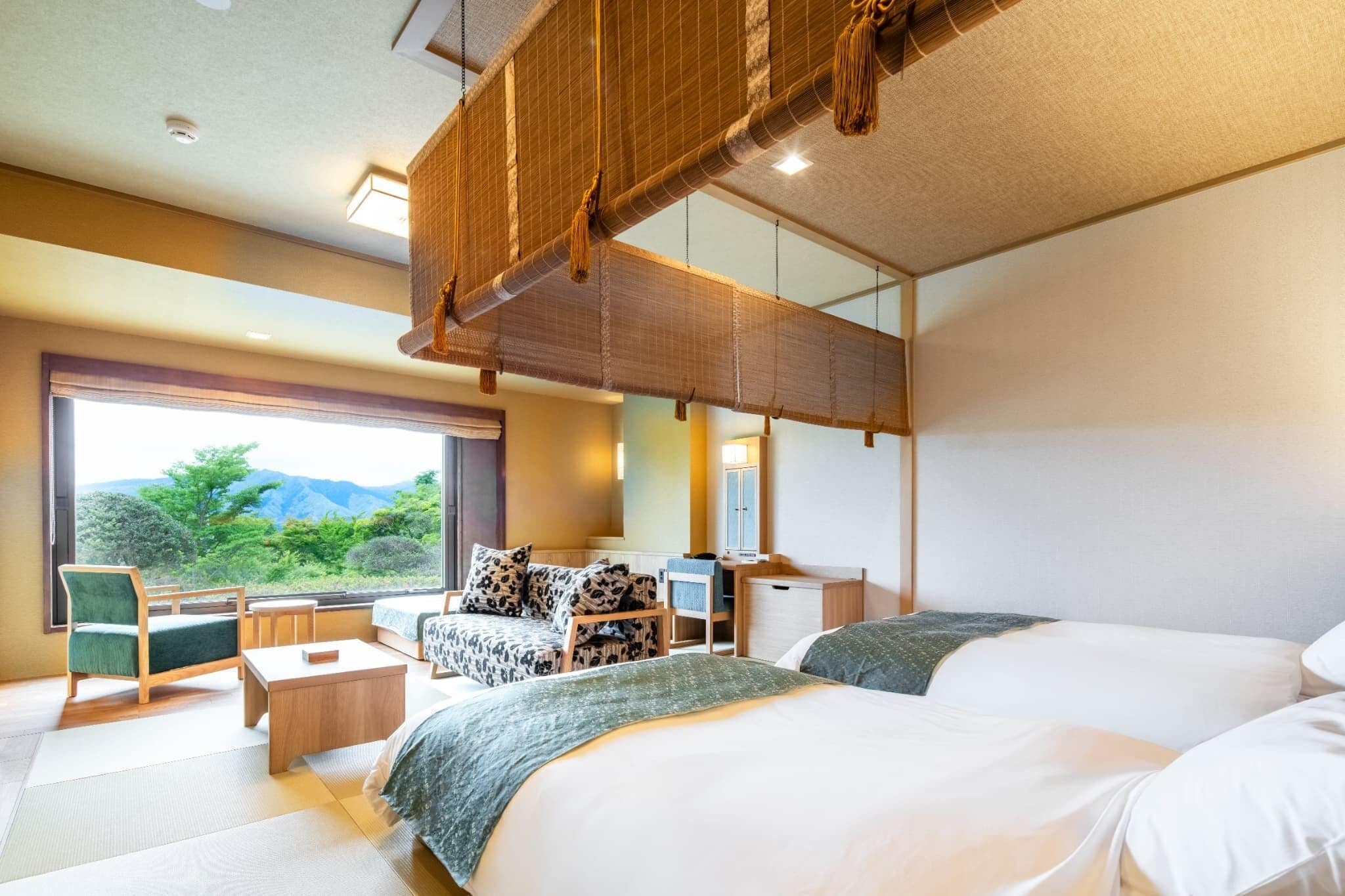 A hotel room with two beds, a desk, a chair, and a large window offering a view of greenery.