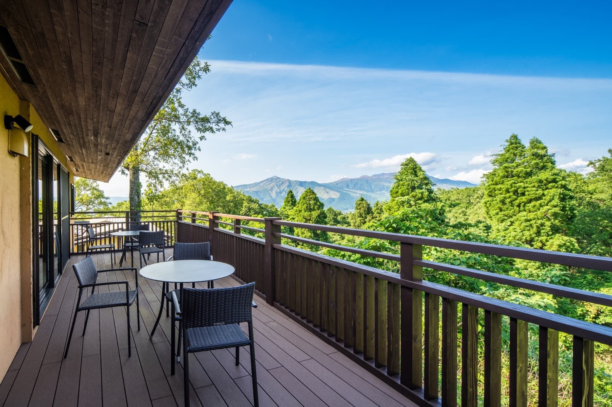 A wooden deck with outdoor seating, a view of green trees and mountains, and a clear sky.