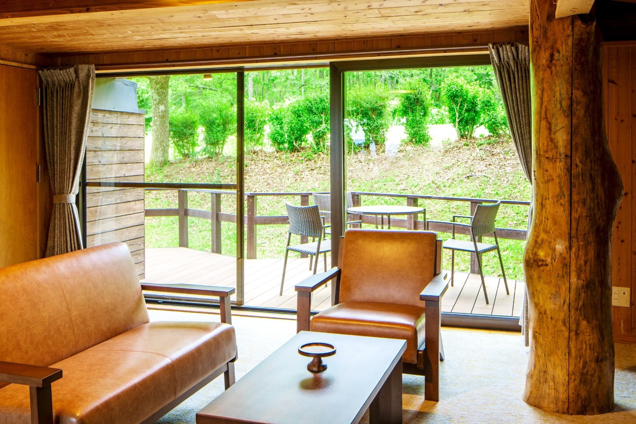 A wooden interior with a sofa, armchair, and table, a large window with curtains, and a view of a green landscape.