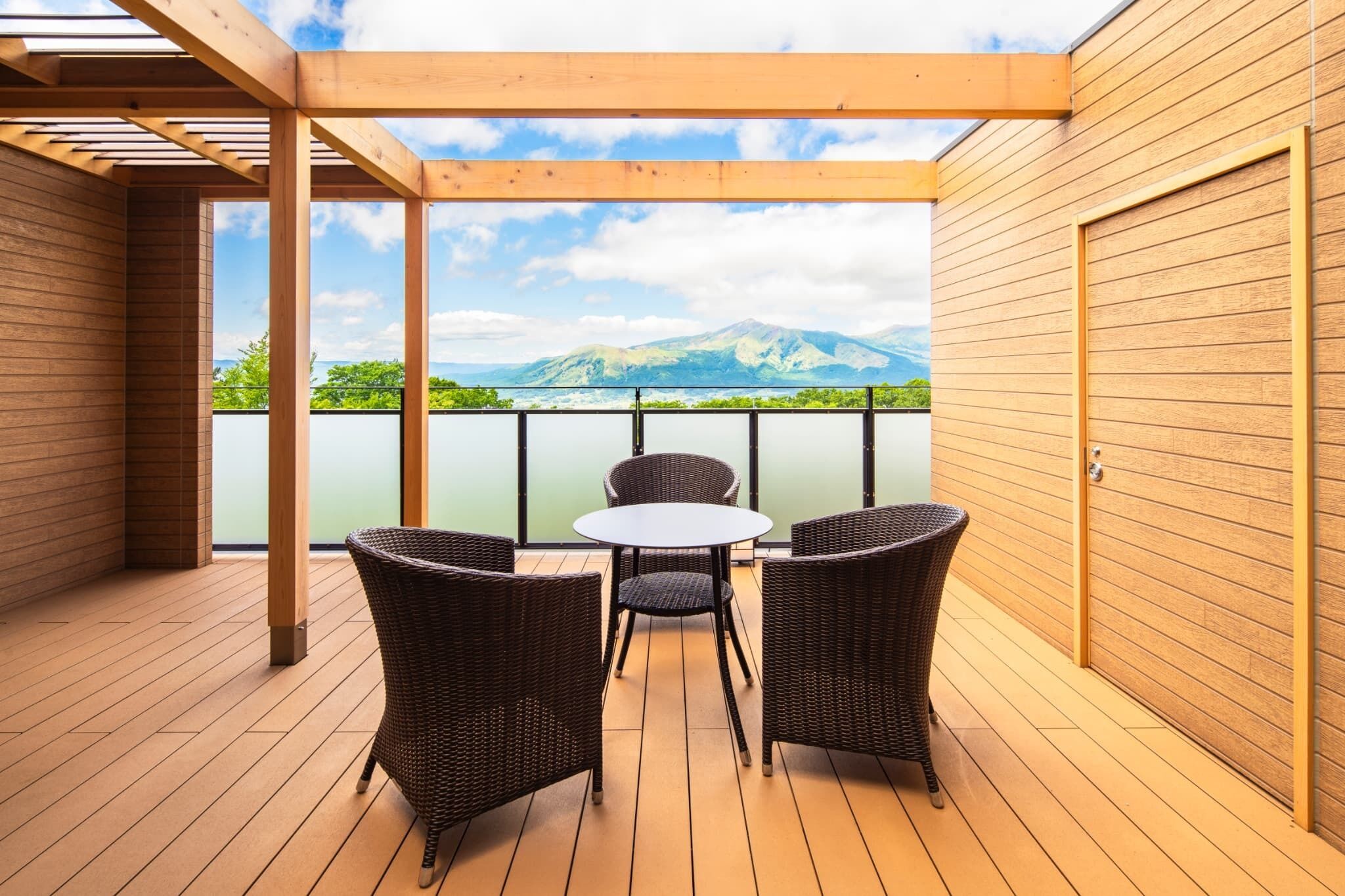 A wooden deck with a table and chairs, overlooking a body of water and mountains.