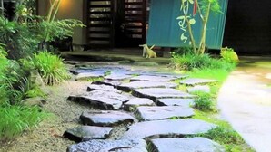Un camino de piedra que conduce a un edificio con una entrada tradicional, rodeado de vegetación y un perro pequeño.